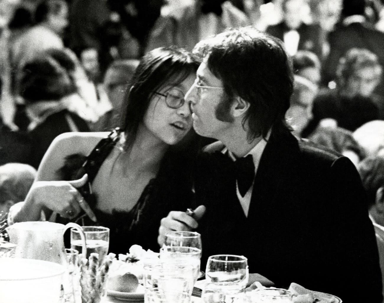 A black and white picture of May Pang and John Lennon sitting at a dinner table during his lost weekend.