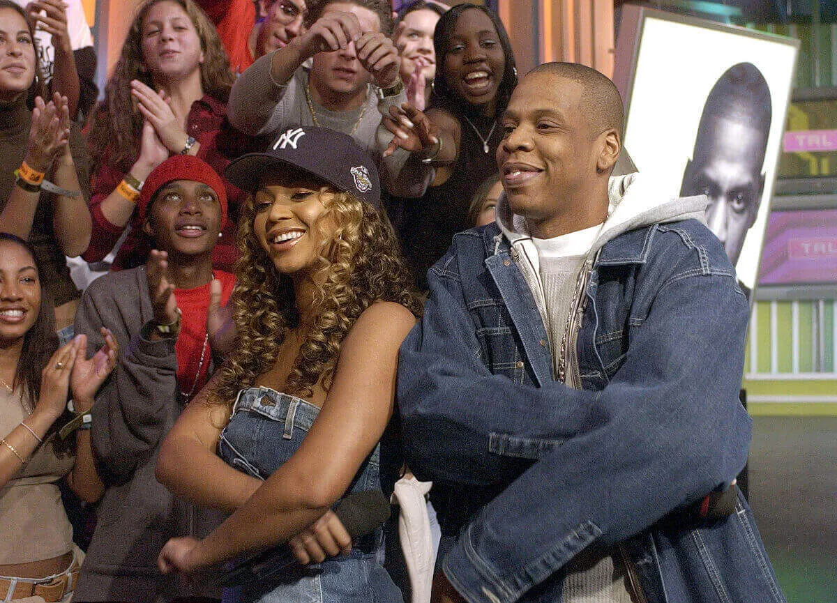 Beyonce and Jay-Z wear denim and stand with their arms folded. She wears a baseball hat.