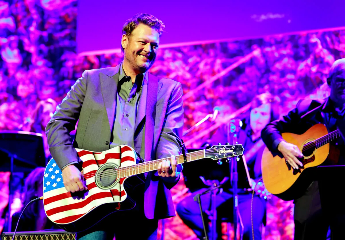 Blake Shelton wears a gray jacket and holds an acoustic guitar decorated like the American flag.