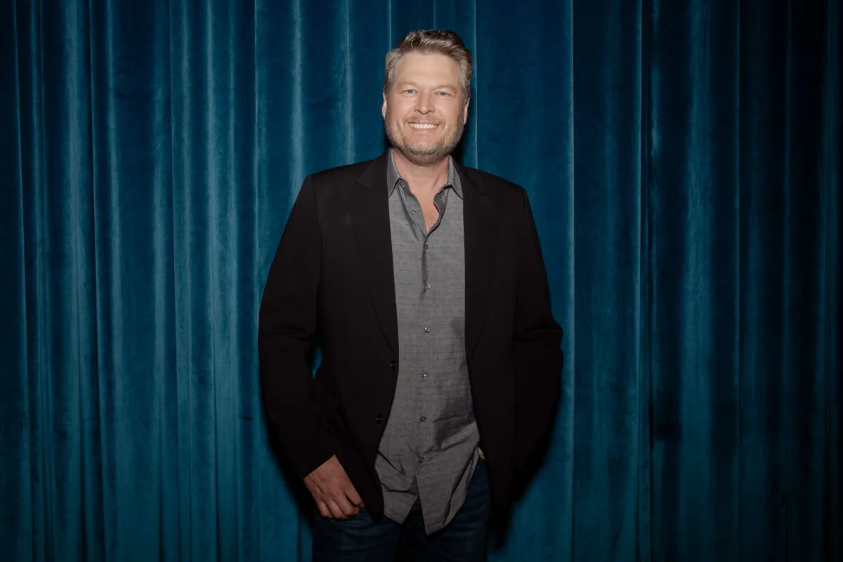 Blake Shelton wears a suit jacket and stands in front of a blue curtain.