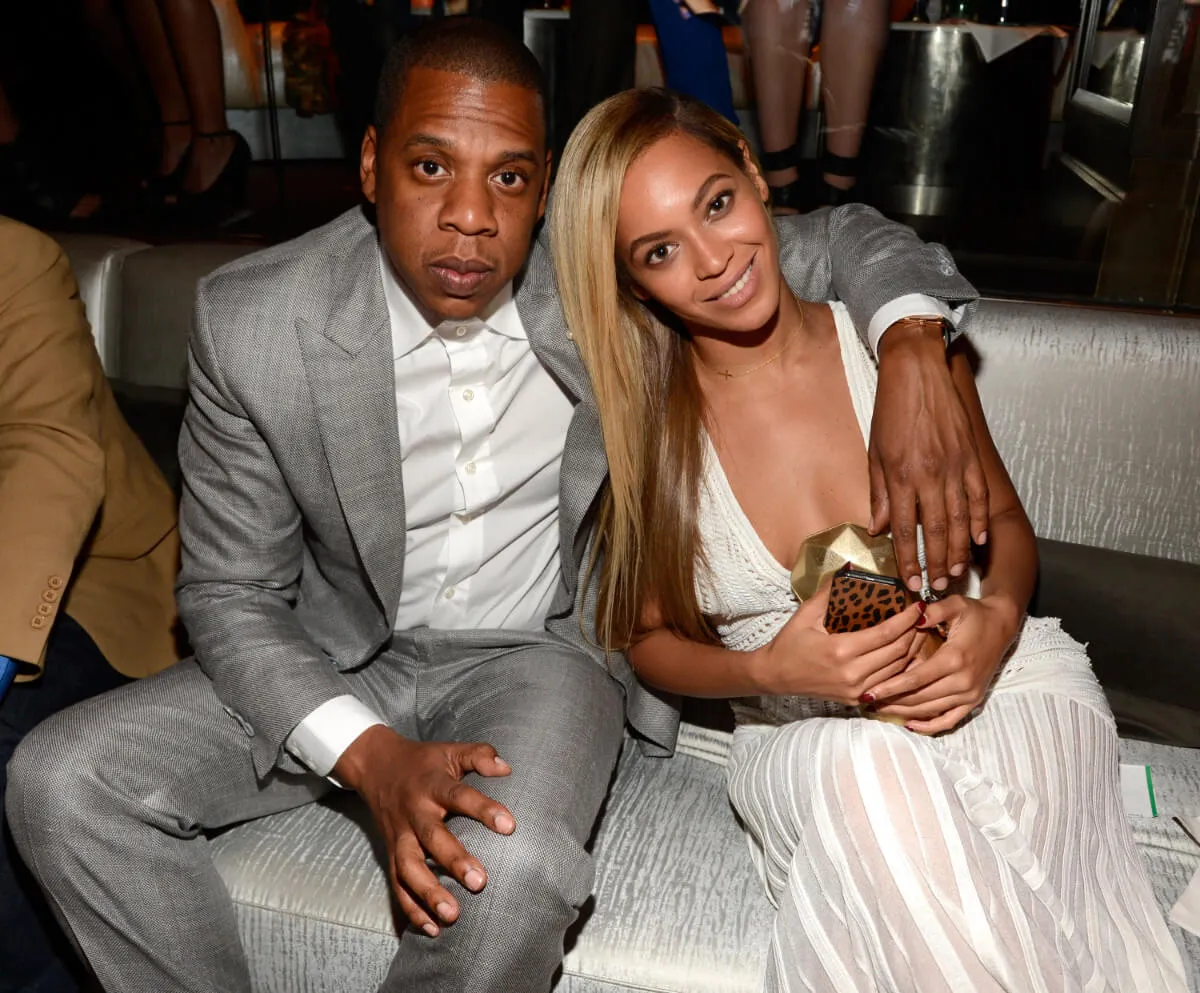 Jay-Z sits with his arm around Beyonce. He wears a gray suit and she wears a white dress.