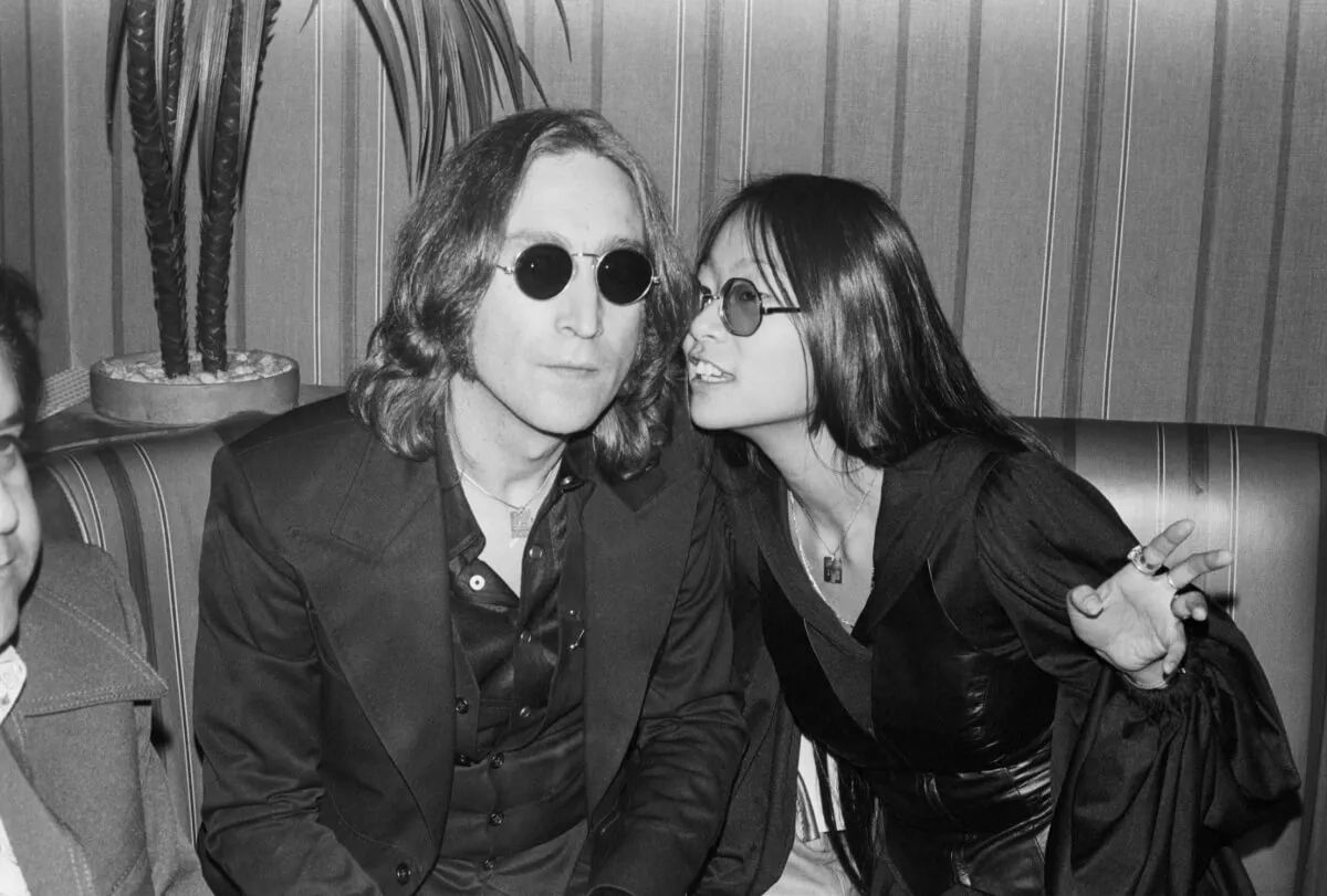 A black and white picture of John Lennon and May Pang sitting in a booth together. They both wear sunglasses and she leans over to talk into his ear.