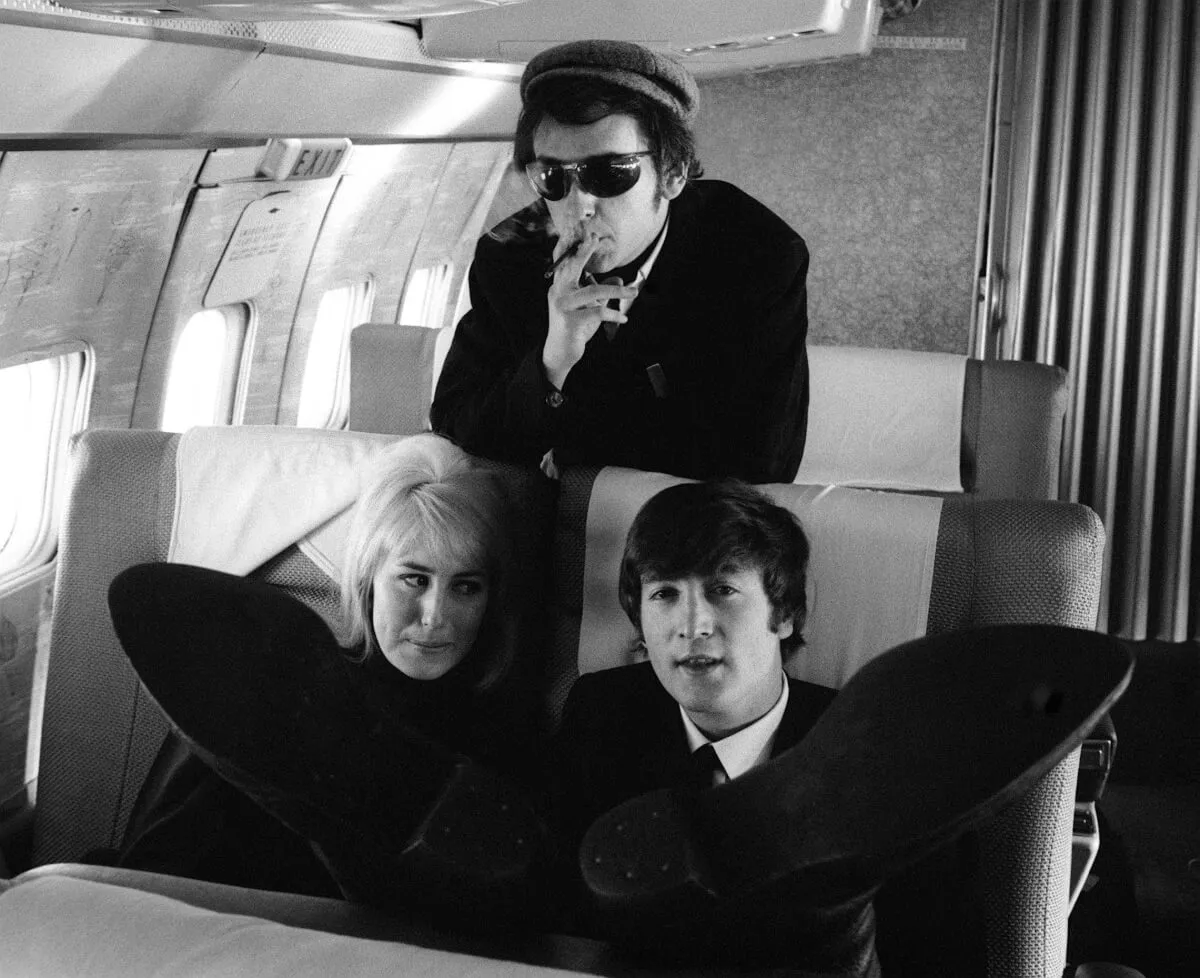 A black and white picture of Cynthia and John Lennon sitting on a plane together. Phil Spector stands behind them in sunglasses. Lennon lifts his feet toward the camera. 