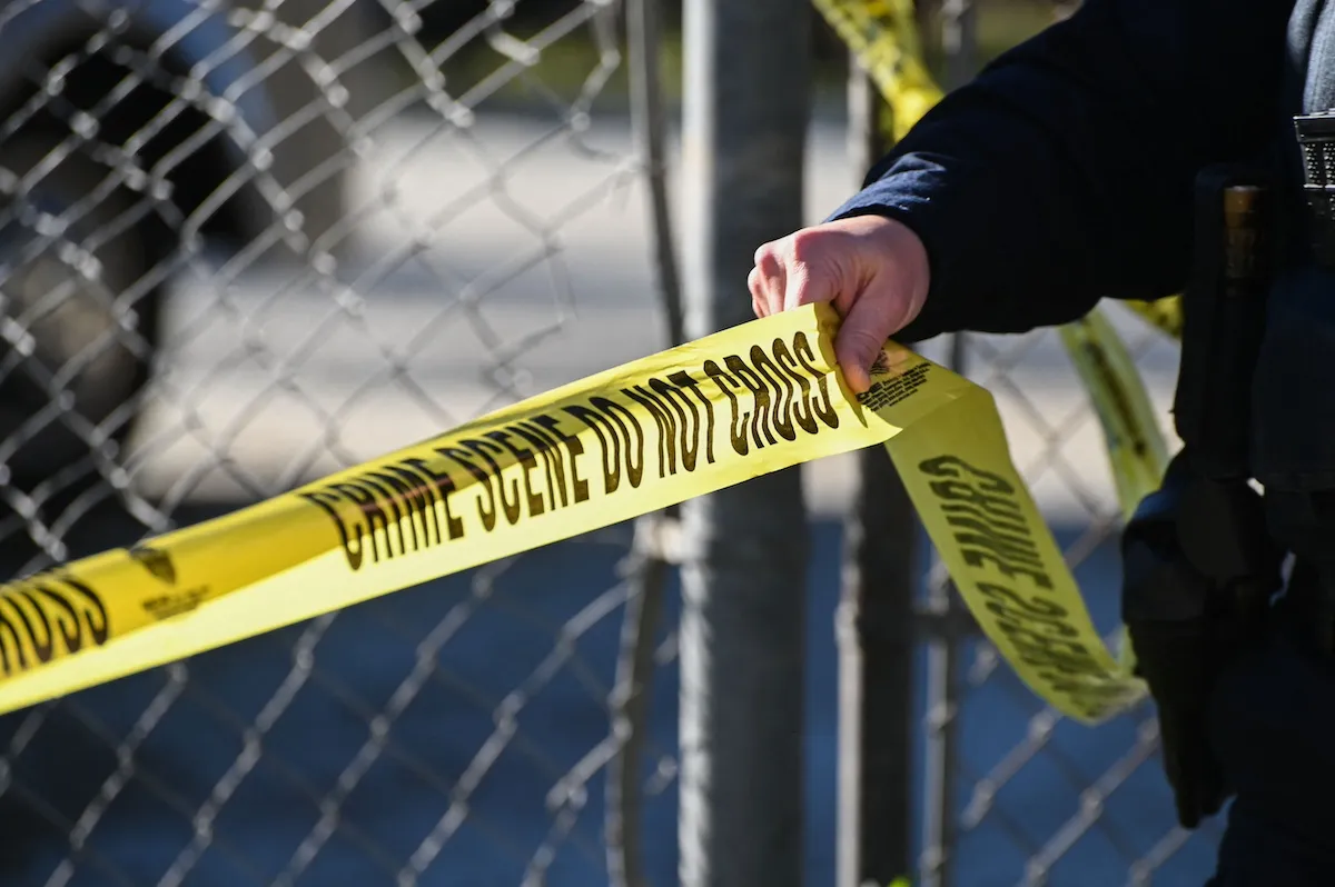 A hand unfurls police tape on a fence at a crime scene