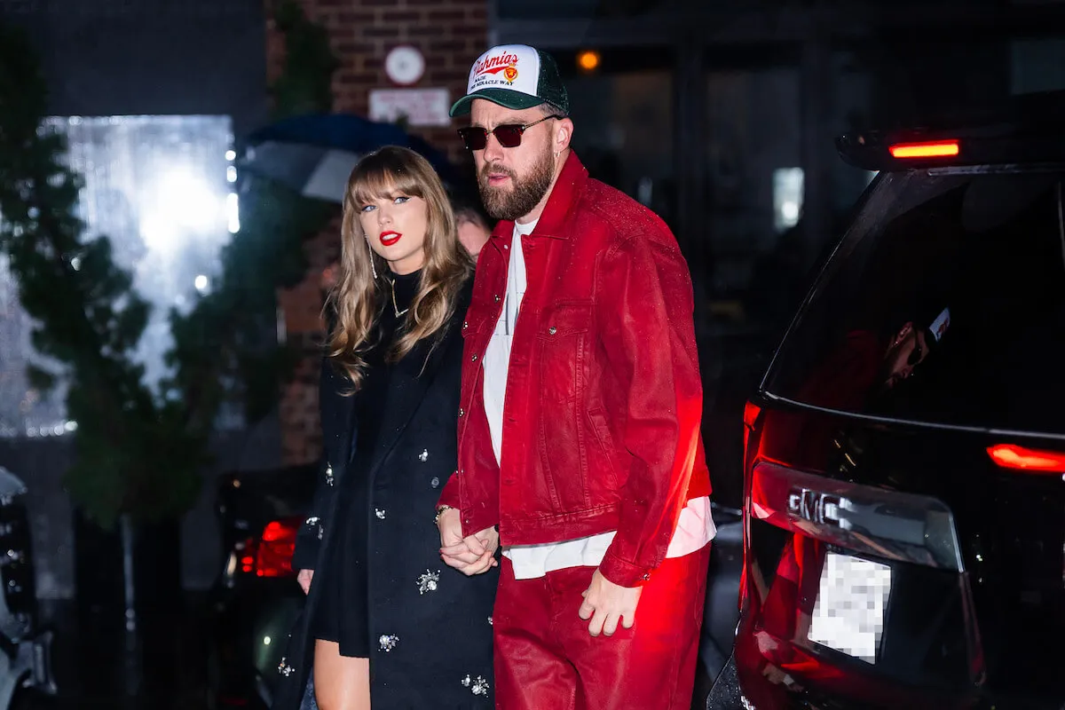 Taylor Swift in a dark dress walking with Travis Kelce at night in New York City, who's dressed in a red jacket and matching pants