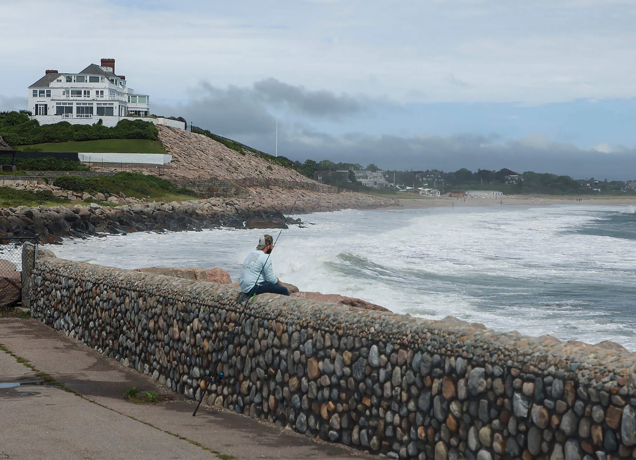 Taylor Swift's Rhode Island home in the background as a fisherman sits on a stone wall on the coast in the foreground