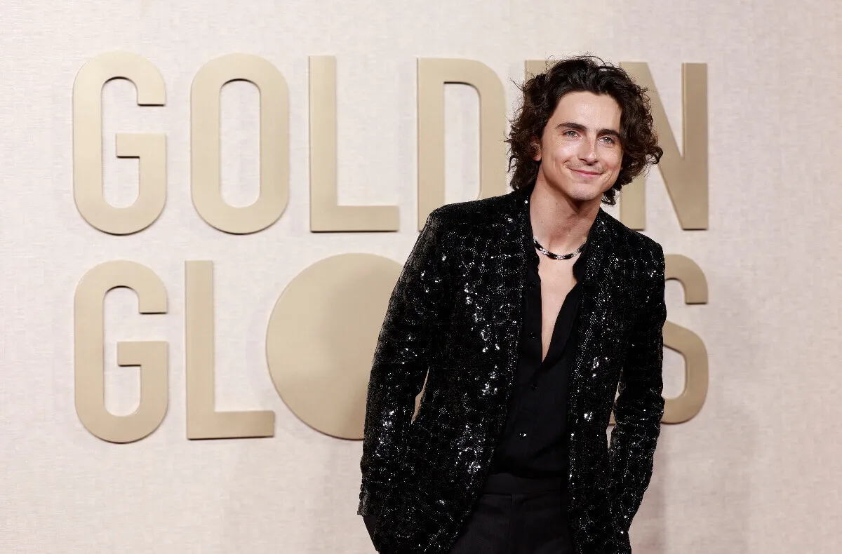 Timothee Chalamet wears a black suit and stands in front of a sign for the Golden Globes.