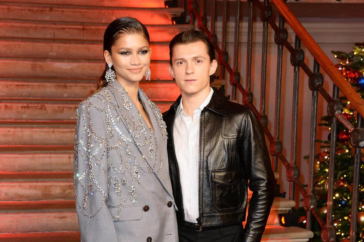 Zendaya and Tom Holland stand in front of a staircase. She wears a silver blazer and he wears a black leather jacket.